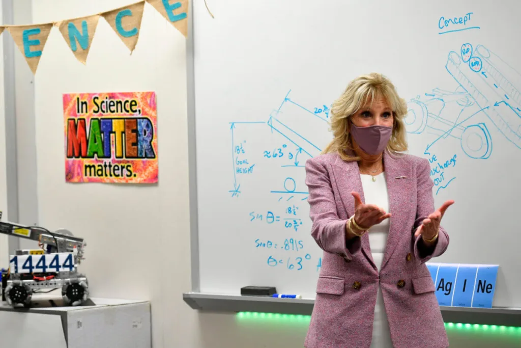 First lady Jill Biden stands in front of a white board in a robotics classroom. She is wearing a white dress, a dusky pink blazer, and a pink mask over her mouth and nose.