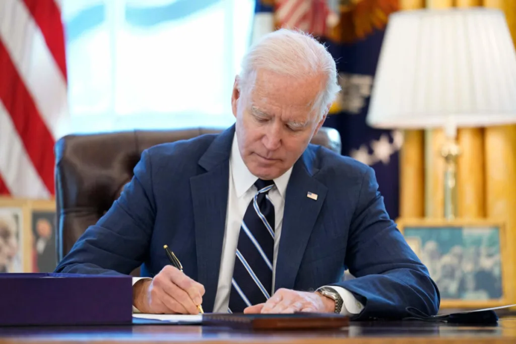 President Joe Biden, wearing a navy blue suit white shirt, and navy blue tie, sits at a dark brown wooden desk as he signs the American Rescue Plan.