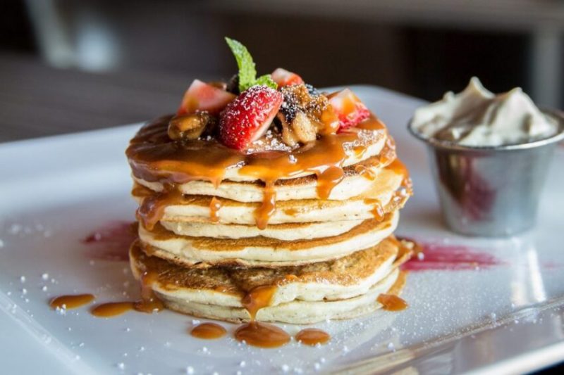 A stack of pancakes covered in strawberries and syrup with a side of butter.
