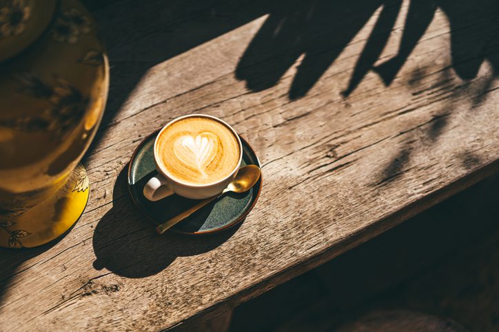 Cup of aromatic cappuccino in ceramic mug in the morning sun. Wooden table and ceramic yellow vase. Happy awakening with comforting milky drink