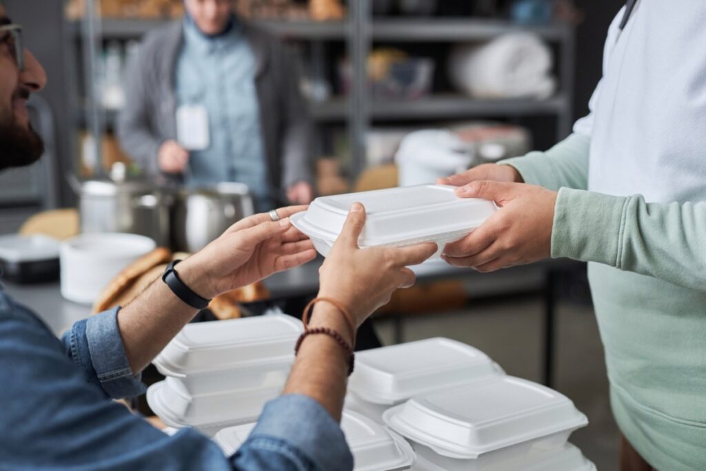 Close up of unrecognizable volunteer giving hot meal in takeaway box to refugees in help center.