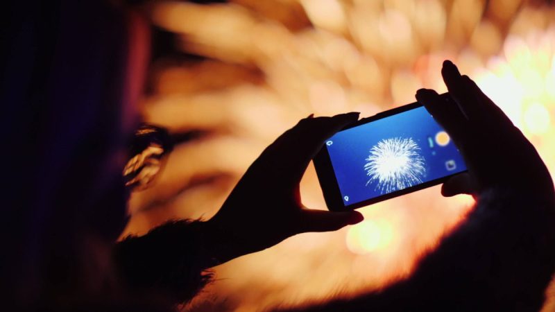 a hand holding a cell phone takes photos of the fireworks going off around them