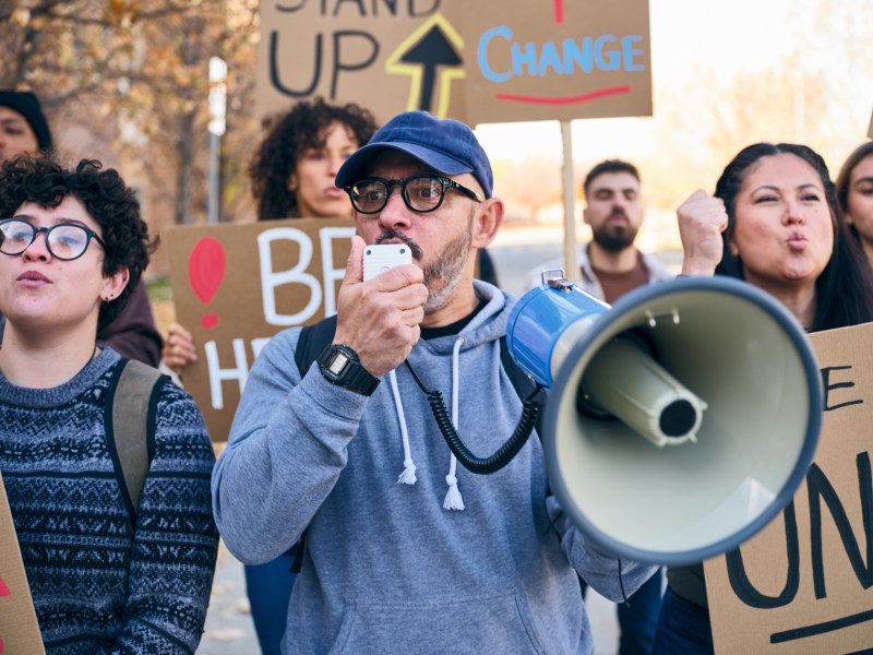A diverse group of adults gathered outdoors holding handmade signs while a man speaks through a megaphone during a positive community rally.