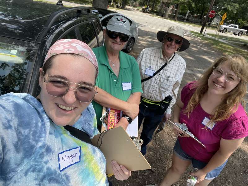Four smiling canvassers wearing name tags and holding clipboards pose for a selfie beside a car on a tree-lined street talking to people about issues including healthcare costs